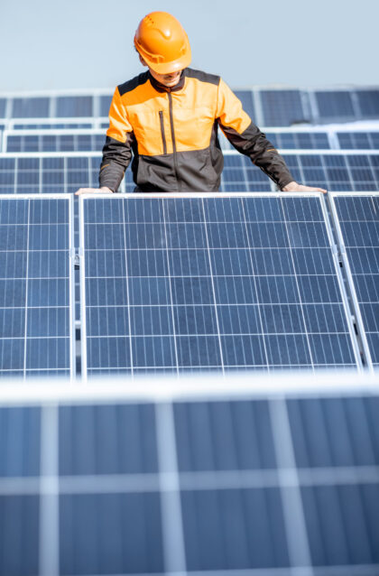 Well-equipped worker in protective orange clothing installing or replacing solar panel on a photovoltaic rooftop plant. Concept of maintenance and installation of solar stations